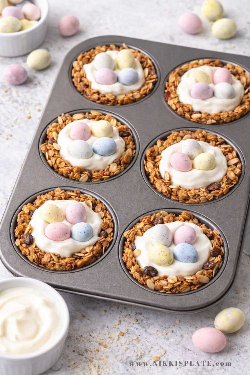 Baked granola nest cups filled with yogurt and topped with pastel mini chocolate eggs in a muffin tin
