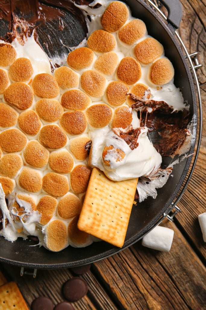 Frying pan with tasty S'mores dip and crackers on wooden background - Easy oven baked s’mores dip recipe; This easy oven baked s’mores dip recipe is perfect for summer BBQs, potlucks and parties. Made with marshmallows, chocolate chips and graham crackers! Crowd pleasing dessert dip!
