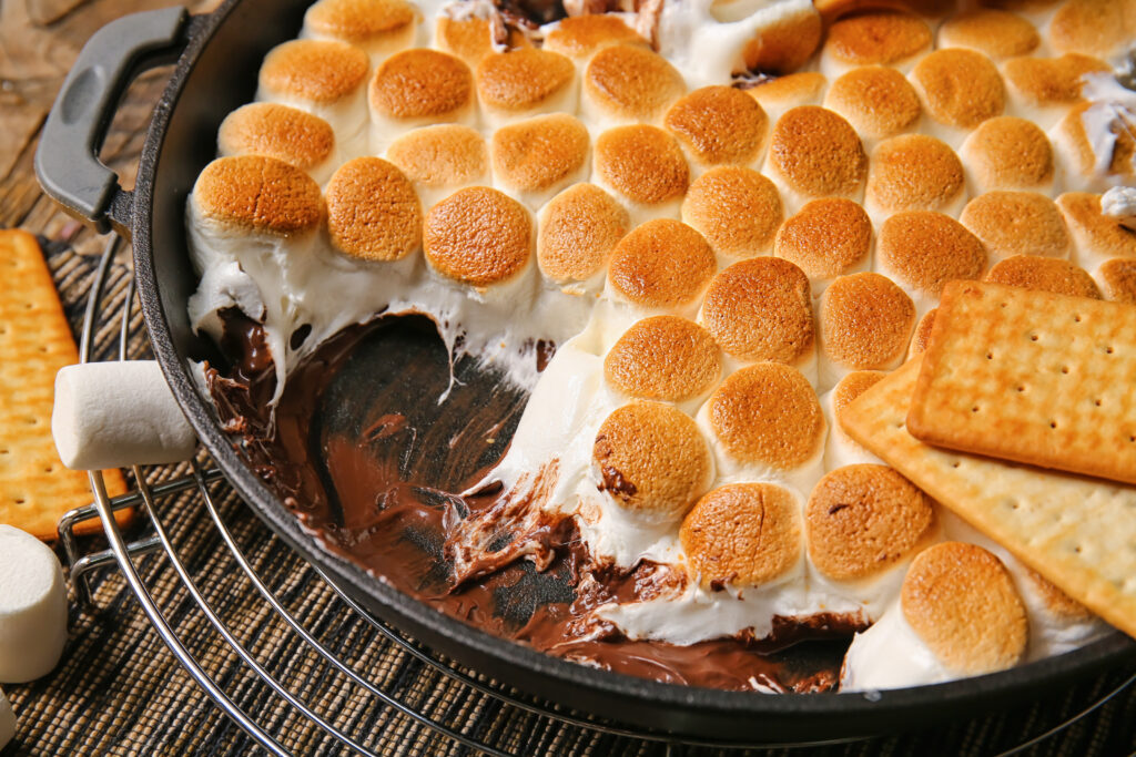 Frying pan with tasty S'mores dip and crackers on wooden table, closeup - Easy oven baked s’mores dip recipe; This easy oven baked s’mores dip recipe is perfect for summer BBQs, potlucks and parties. Made with marshmallows, chocolate chips and graham crackers! Crowd pleasing dessert dip!