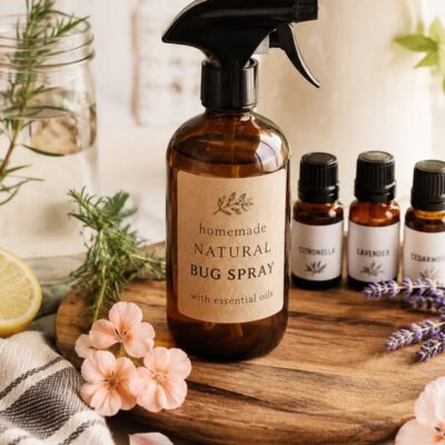 Homemade natural bug spray in an amber glass bottle with citronella, lavender, cedarwood, rosemary, and fresh flowers on a warm farmhouse-style table.