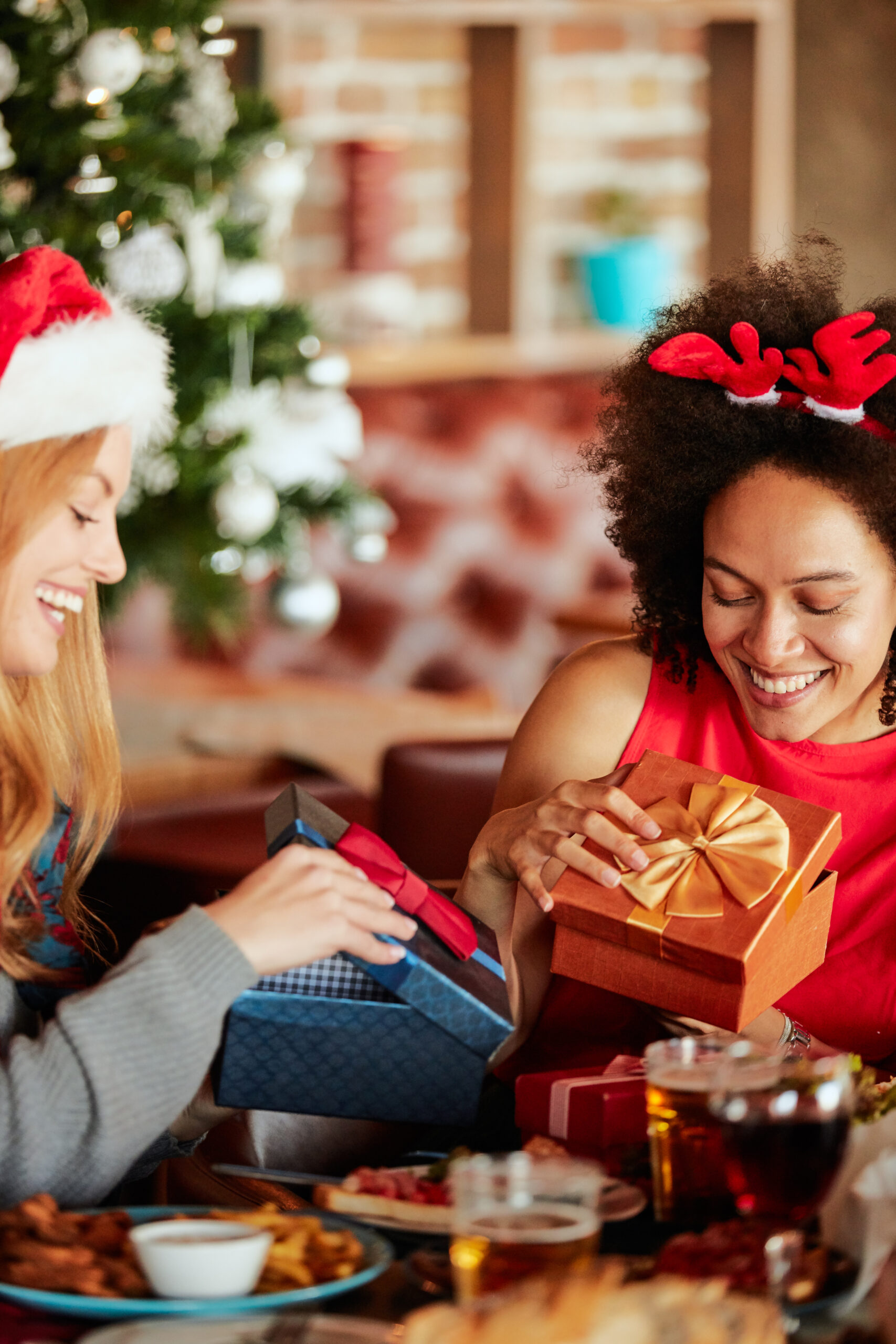 Friends giving gifts to each other while sitting at table. In background Christmas tree. Christmas holidays concept.