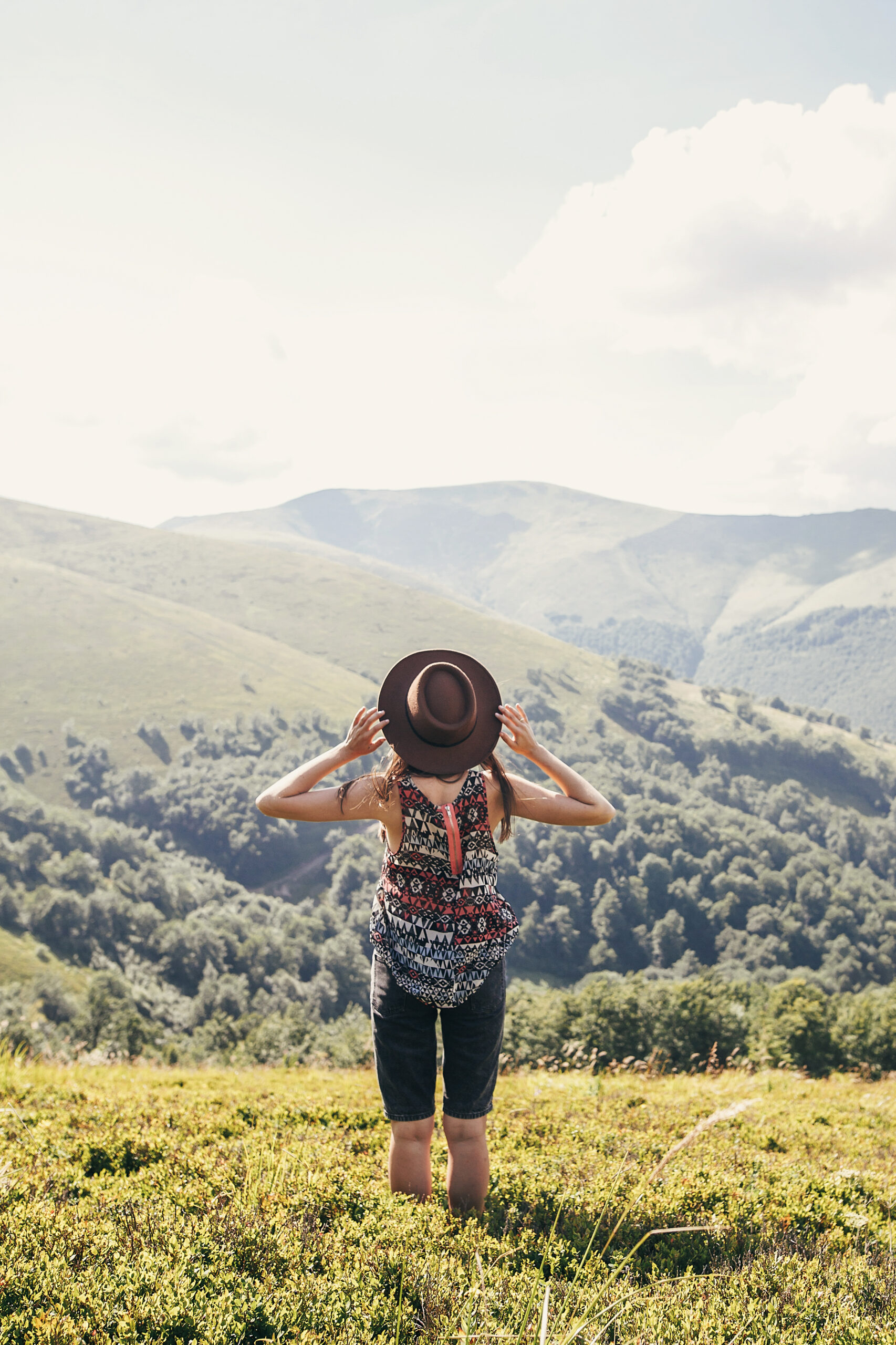 stylish traveler girl in hat looking at sunny mountains. summer vacation. travel and wanderlust concept. space for text. back view. woman traveling. amazing atmospheric moment - best travel hacks for saving money