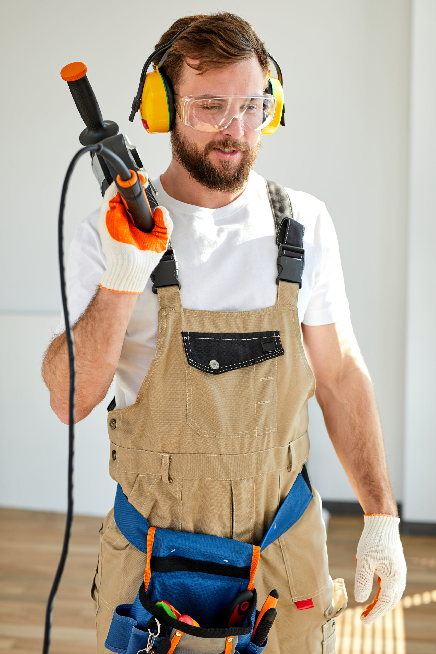 Young caucasian man holding power drill standing in front of white wall indoors,posing at camera, wearing overalls, headset and safety glasses. copy space, concept for home DIY - Safety Equipment You need when Doing DIY in the Home
