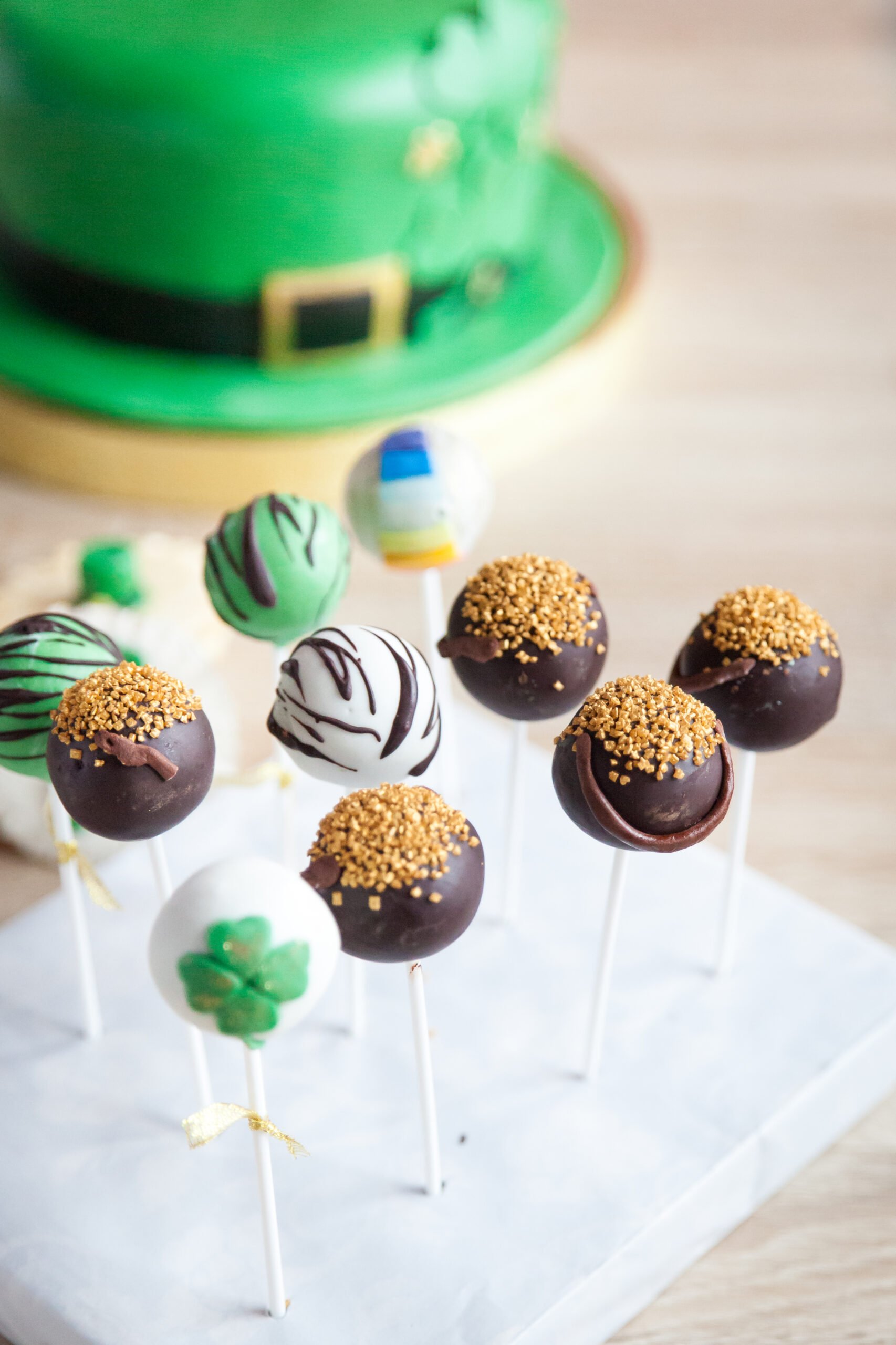 Funny cake pops for St Patrick's Day and green cake in the shape of a St. Patrick's hat in the background. First birthday party sweets - Top 10 St. Patrick's Day Dessert Recipes; St. Patrick's day recipes for desserts along with other green recipes for this Irish holiday!