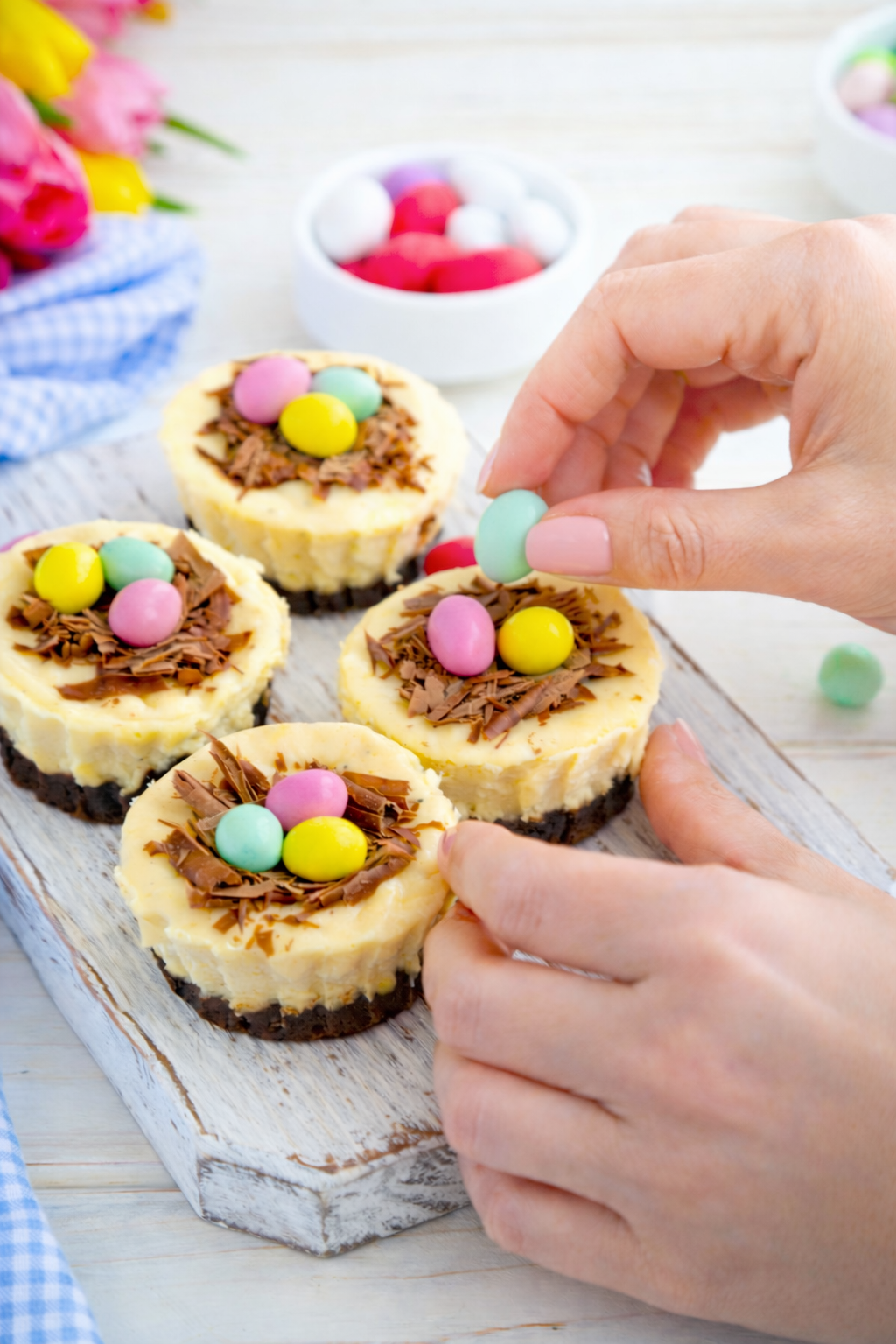 Woman's hands decorating Easter brownie cheesecake birds nests with pastel candy eggs on a rustic wooden board.