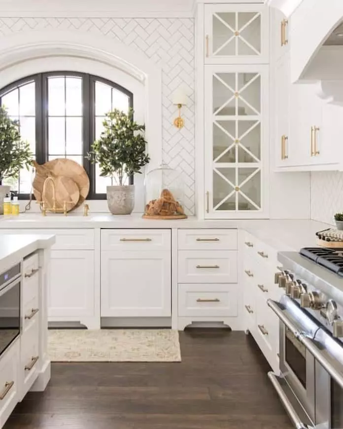A window with an arched top and black frame is surrounded by white herringbone tiles. A gold sconce with a white shade matches the gold handles on the cabinets, which host stainless steel appliances. Plants and cutting boards are arranged in front of the window.