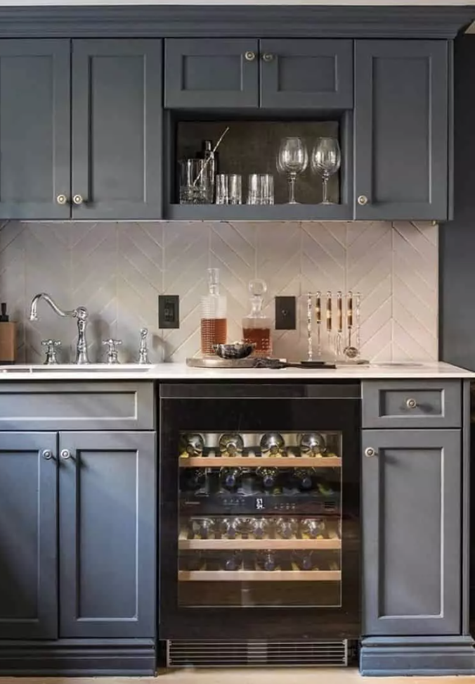 Dark gray shaker cabinets are paired with a gray subway tile herringbone pattern kitchen backsplash. A white countertop and black glass front wine fridge tops the cabinets.