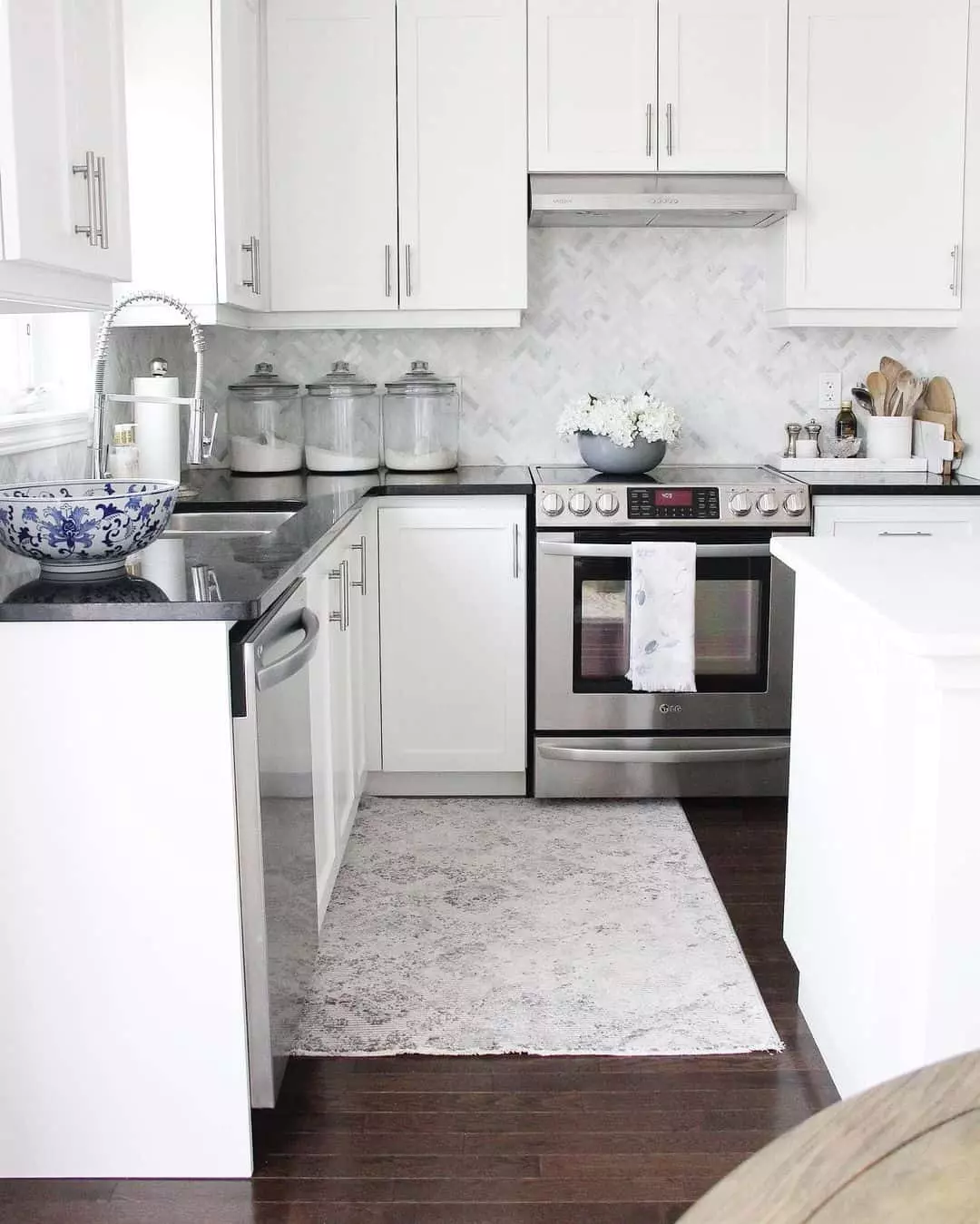 Nickel pulls add a touch of elegance to this L-shaped kitchen. A stainless steel range and range hood are fitted between the shaker cabinets in front of a white and gray herringbone tile backsplash.