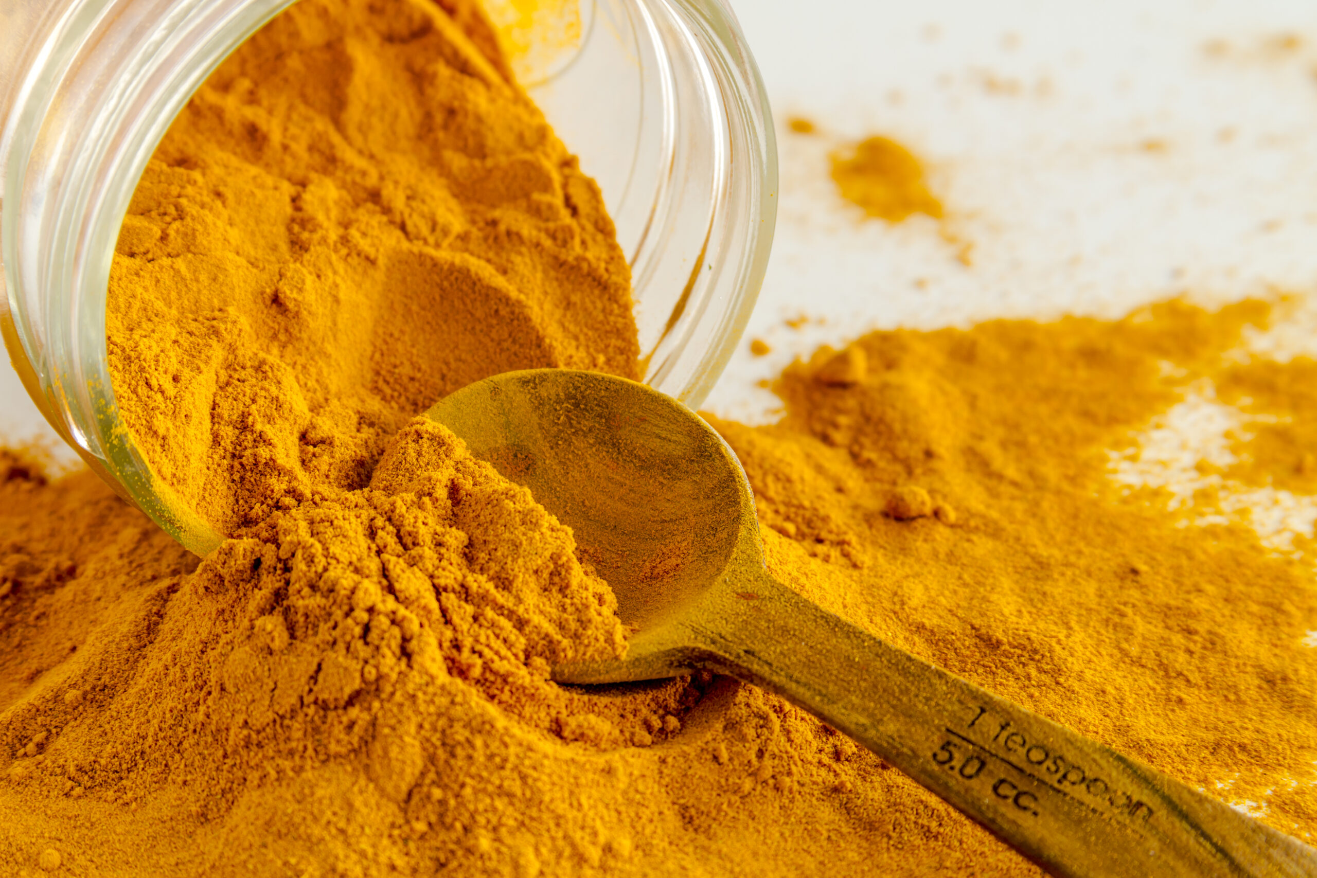 Close up of organic turmeric (curcuma) powder spilling out of glass jar with measuring spoon on white background