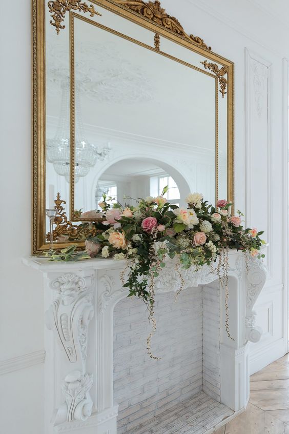 Mirror above fireplace ideas; A magnificent large brass mirror takes center stage above a beautifully adorned fireplace. The mirror's lavish frame radiates a sense of opulence, while its oversized dimensions command attention. Nestled on the mantelpiece, an exquisite floral arrangement bursts with vibrant colors, creating a harmonious and eye-catching contrast against the shimmering brass.