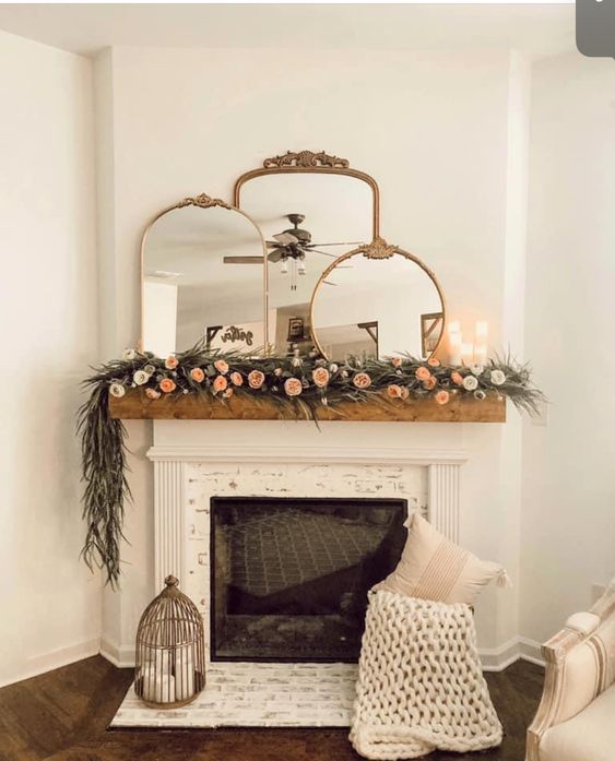Mirror above fireplace ideas; A stunning white fireplace adorned with three layered mirrors and a gorgeous floral garland draped across the mantel. The chic white fireplace provides a sleek, modern touch to the space, while the mirrors add a sense of depth and reflect the natural beauty around the room, illuminating every corner with a dazzling glow. The exquisite floral garland is the perfect finishing touch, adding a pop of color and texture that draws the eye in and elevates the atmosphere to one of pure charm and serenity.