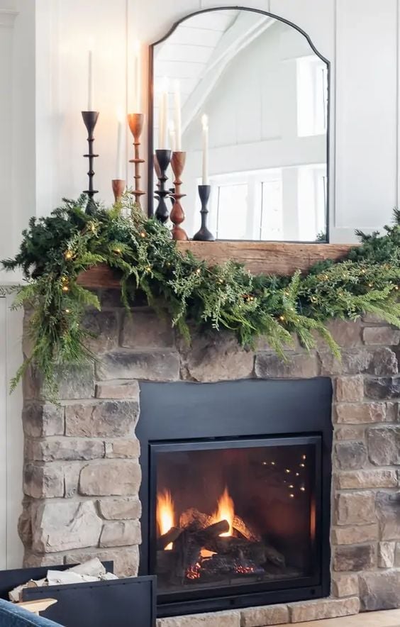 Mirror above fireplace ideas; This festive photo showcases a stunning Christmas mantel that has been adorned with a beautiful mirror, garland, and candlesticks. The mirror adds depth and reflection to the space, creating a captivating visual display. The lush green garland and flickering candlesticks bring warmth and coziness to the decor, making the mantel a welcoming and inviting focal point in the room.