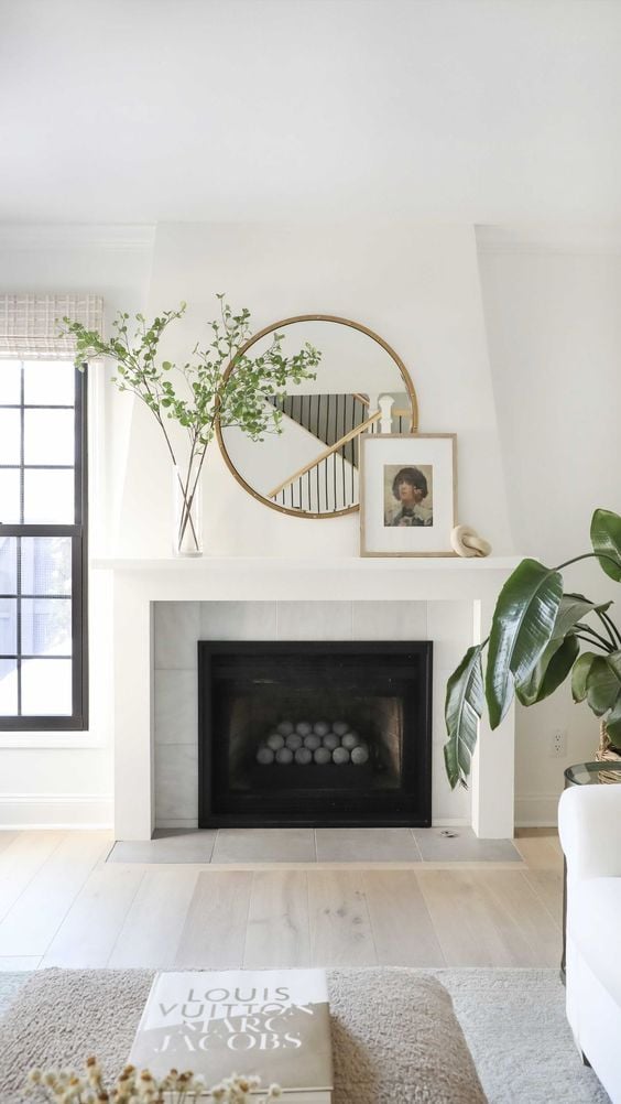 Mirror above fireplace ideas: A small round mirror is artfully layered on the fireplace mantle, surrounded by delicate greenery and a picture frame. 