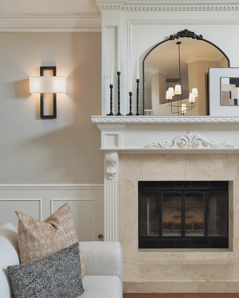 Arched black-framed mirror styled above a traditional white fireplace mantel with tall black candlesticks and layered artwork in a sophisticated living room.