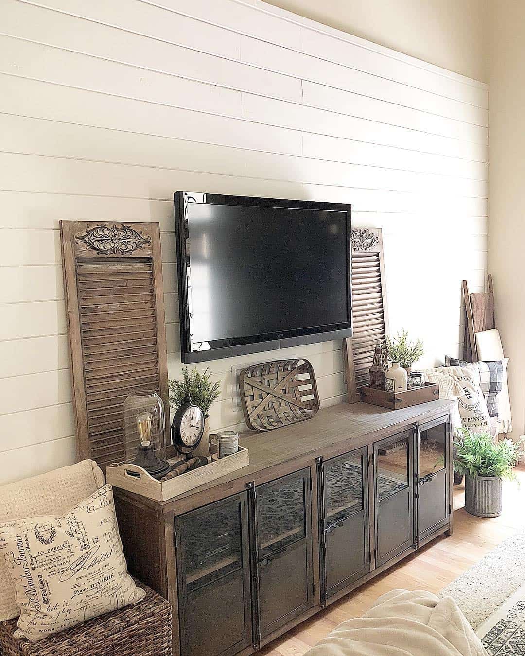 farmhouse TV stand ideas; In this photo, the rustic textures of the wooden shutters provide a striking contrast against the pristine white shiplap walls while effortlessly complementing the TV stand. These shutters also create a captivating backdrop for the charming wooden tray vignettes adorned with vintage décor. Additionally, the inclusion of a wicker basket in the center adds a tactile element that harmonizes and completes the overall composition.