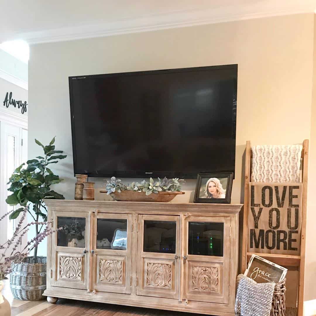 farmhouse TV stand ideas; Enhancing the space between the TV and the TV stand is a remarkable long wooden trough brimming with lush leaves. The trough is artfully adorned with antique jars and a captivating picture frame, creating a seamless and visually pleasing display. Its rich wood finish perfectly complements the TV stand, resulting in a cohesive and harmonious aesthetic.