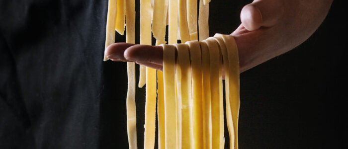 Culinary Escapades - Fresh raw uncooked homemade pasta tagliatelle in man's hands over black apron as background.