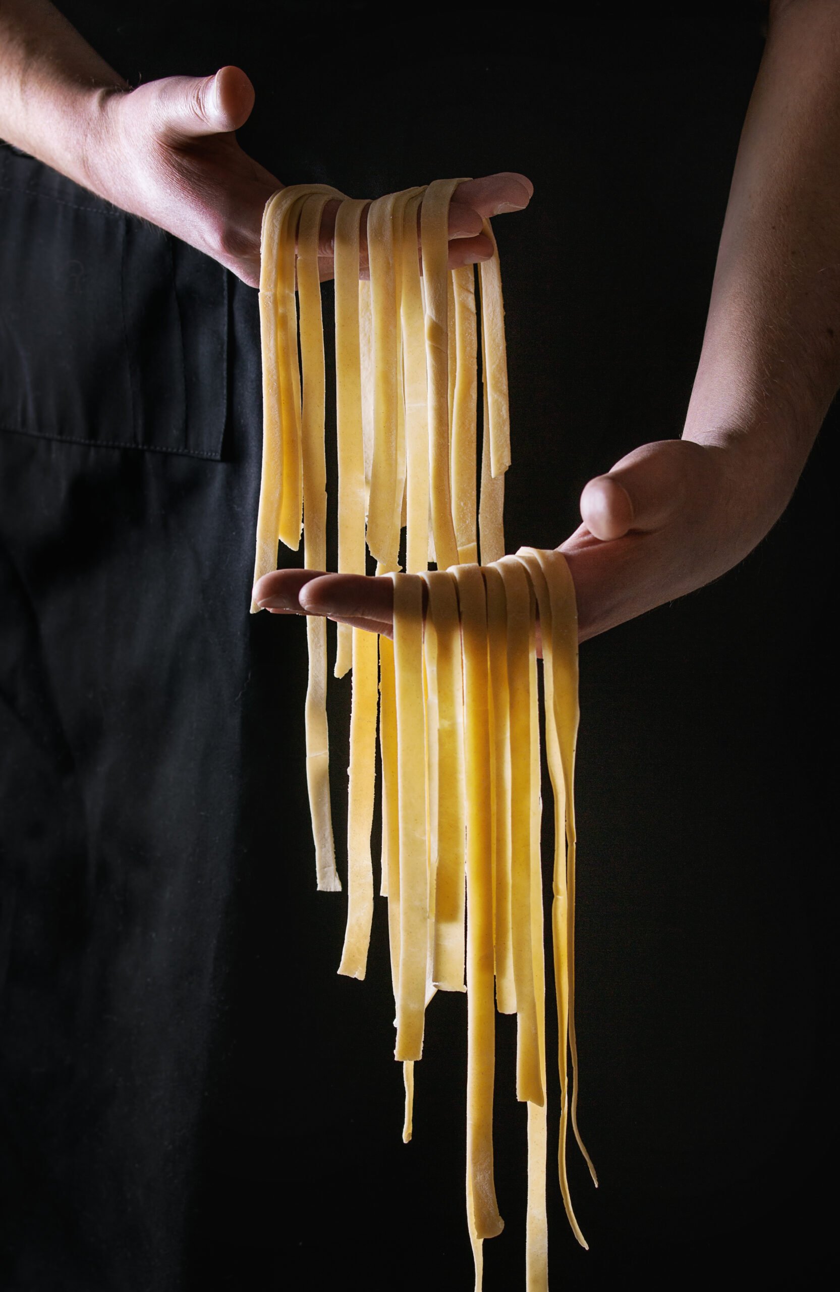 Culinary Escapades - Fresh raw uncooked homemade pasta tagliatelle in man's hands over black apron as background.
