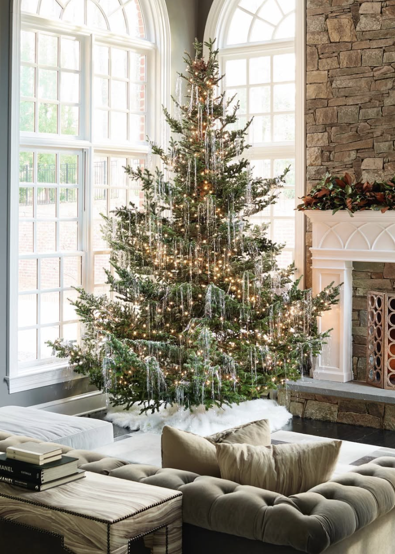 Modern minimalist tinsel Christmas tree with silver tinsel and warm white lights in a bright living room with stone fireplace and large windows.