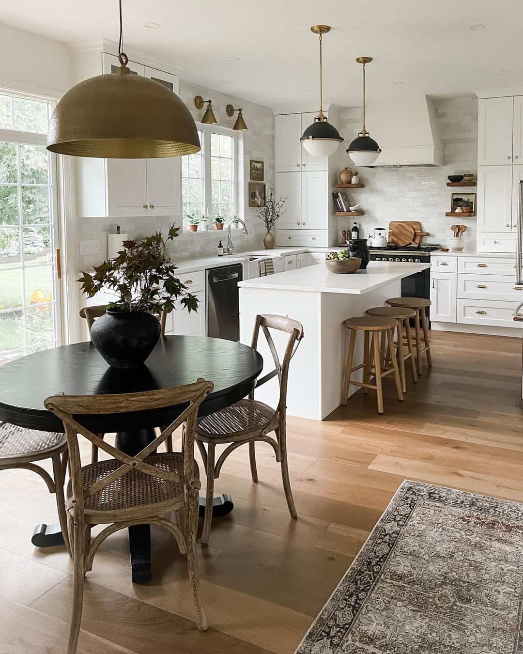 This inviting L-shaped kitchen, enhanced by a center island, is elegantly outfitted with white cabinets adorned with sophisticated gold hardware, all set against pristine white countertops. Suspended above the modern stainless steel stove is a sleek white hood, adding a touch of refinement. Illuminating the scene, a pair of pendulum lamps in a crisp black and white design cast a welcoming glow onto the island's white countertop. Complementing the arrangement, three wooden stools stand upon the inviting warmth of the wooden flooring, creating the perfect spot for gathering and making memories.