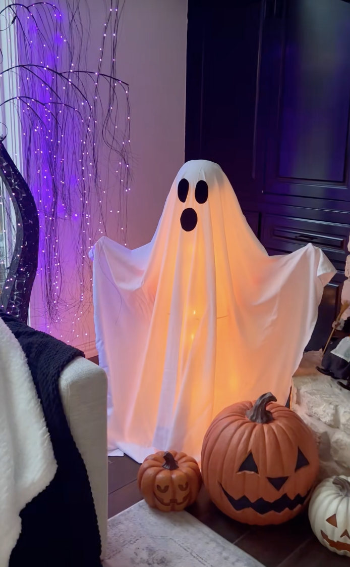 Glowing DIY sheet ghost with black felt eyes, illuminated from inside with string lights and surrounded by pumpkins in a cozy living room corner.