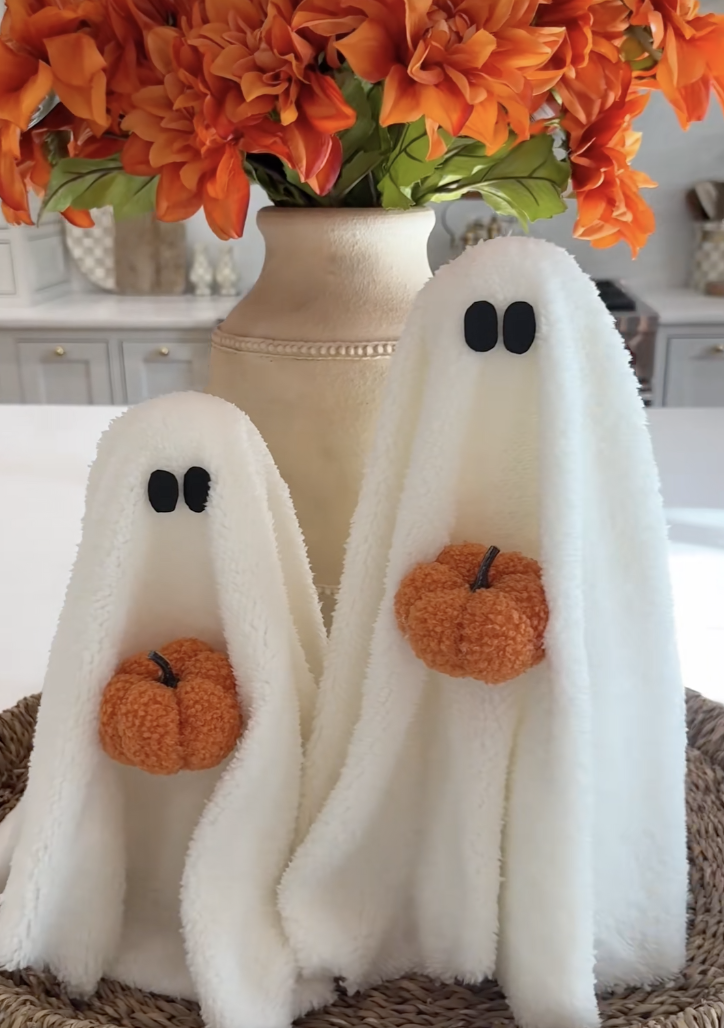 Two DIY towel ghosts holding small orange pom-pom pumpkins, displayed in a woven tray with a vase of bright fall flowers.