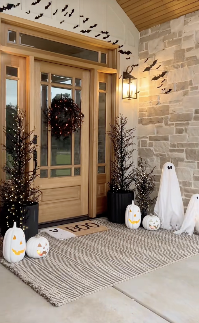 Front porch decorated with glowing white pumpkins, mini standing ghosts, lighted black trees, and a