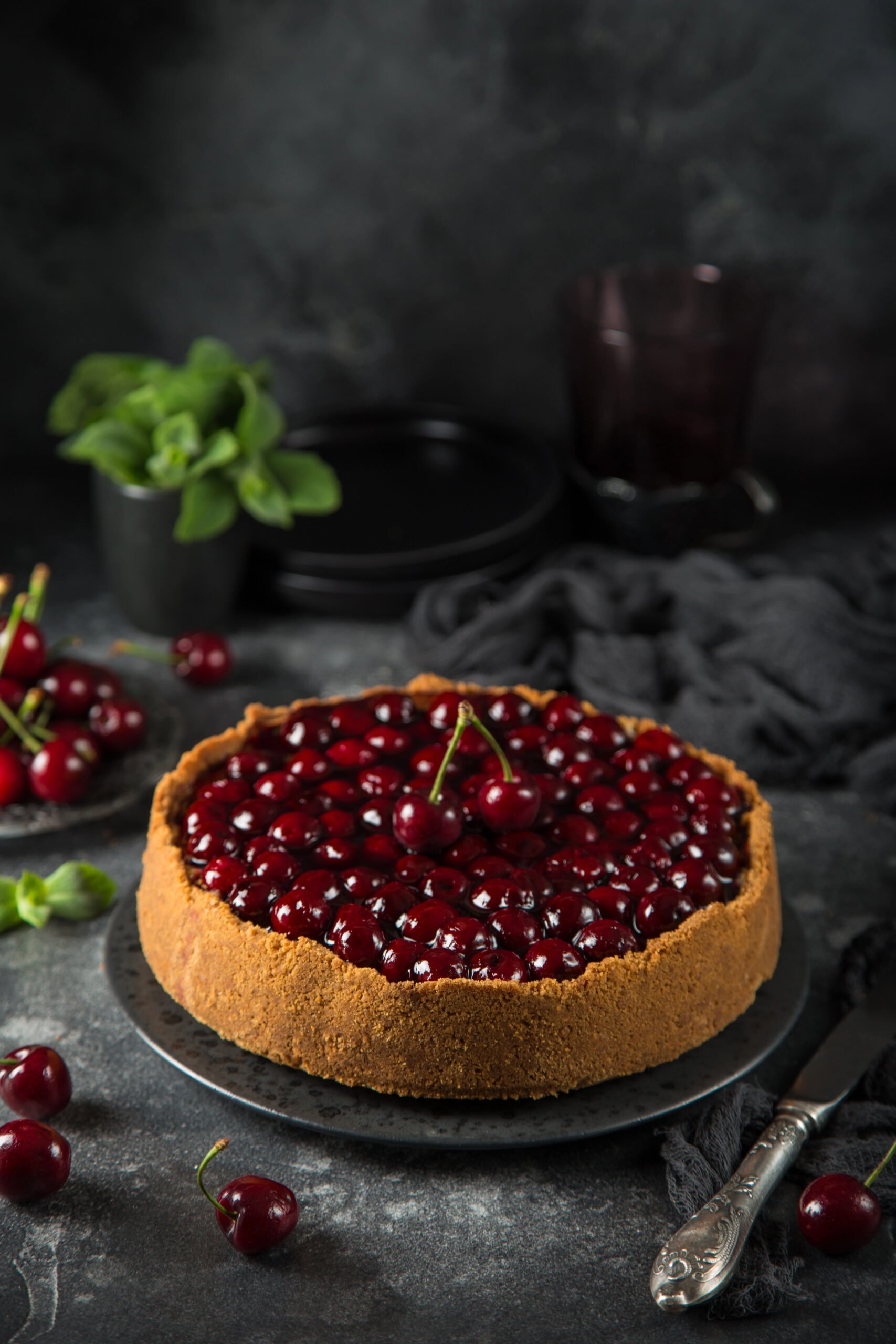 Whole cherry cheesecake with graham cracker crust topped with glossy cherries, styled with fresh mint and cherries on a dark background.