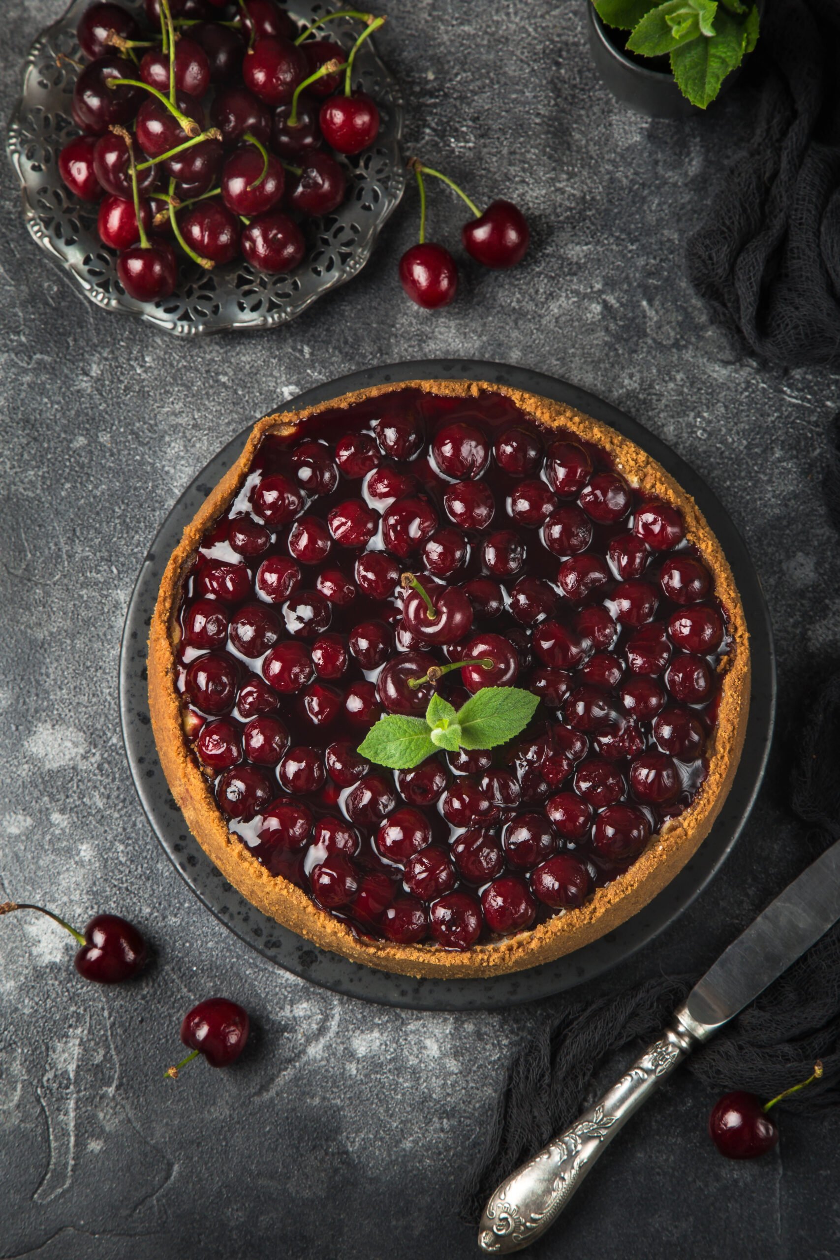 Top view of cherry cheesecake on a black plate, garnished with mint leaves and surrounded by fresh cherries and mint.