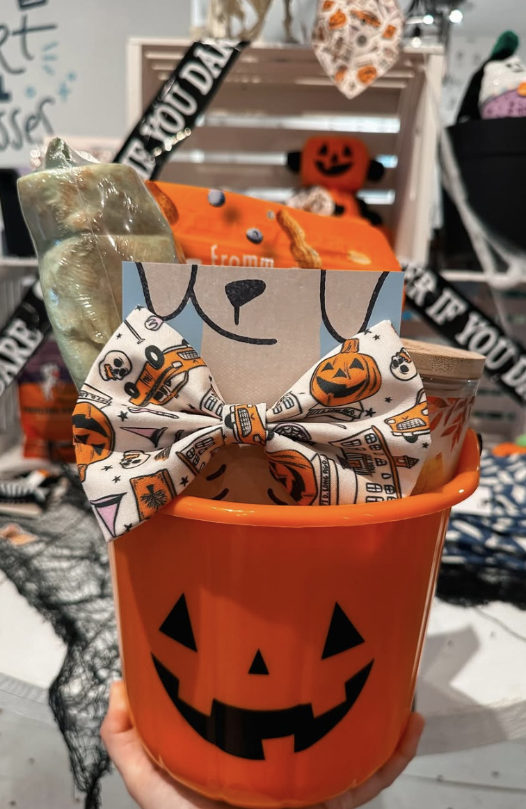 Orange jack-o’-lantern Halloween boo basket for dogs featuring a festive pumpkin-patterned bow tie, treats, and chew toys