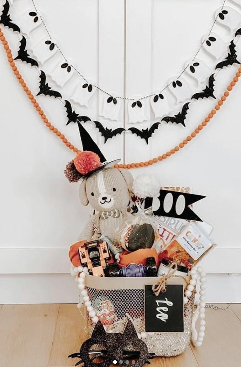Neutral Halloween boo basket with a plush dog toy, festive treats, and black cat mask, displayed under ghost and bat garland decor