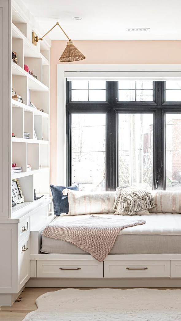 Bright built-in reading nook with blush pink walls, black window frame, and layered pillows