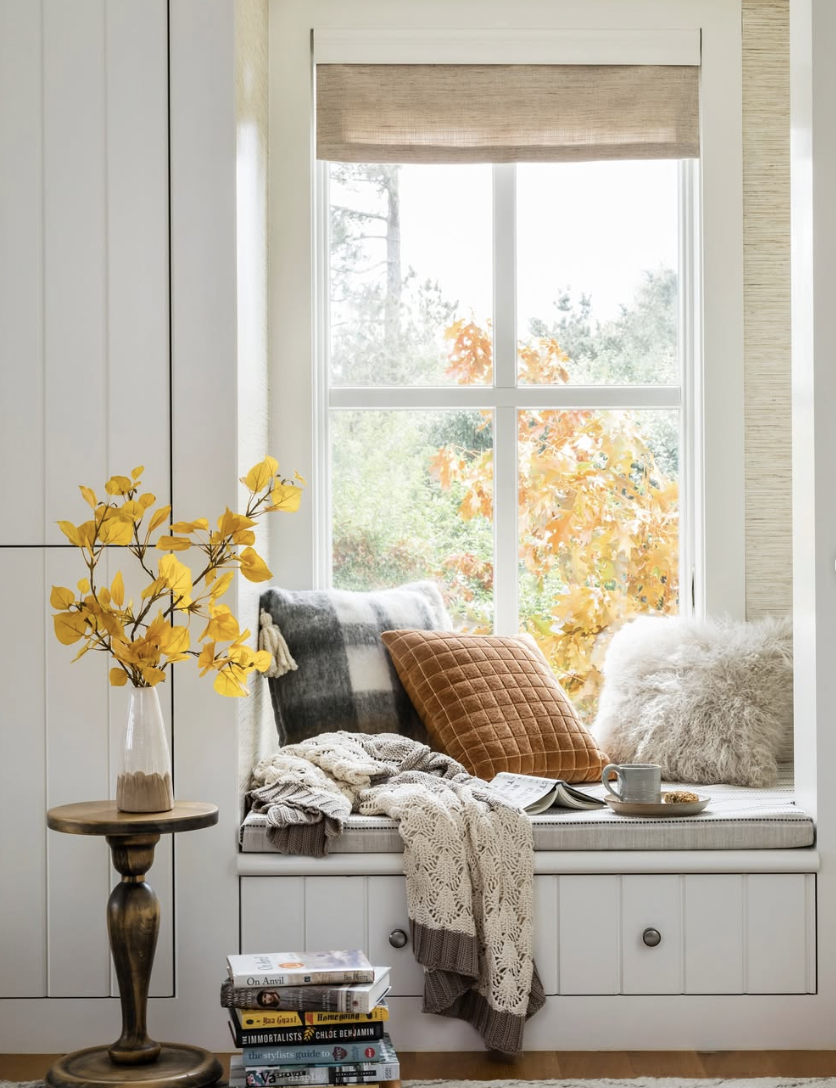 Window seat reading nook with fall foliage view, layered blankets, and warm-toned pillows