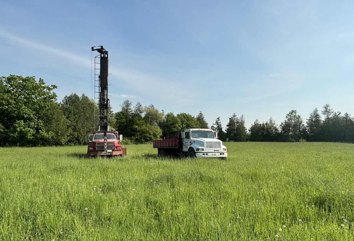 Front view of drilling trucks on a rural property preparing to drill a water well in a grassy field with forest backdrop.