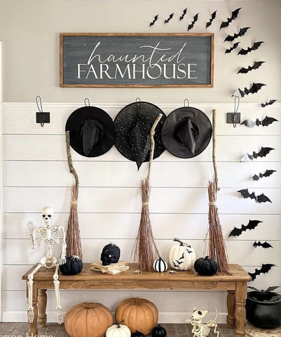 Farmhouse entryway with “Haunted Farmhouse” sign, bat wall decals, witch hats and brooms, skeletons, and black and white pumpkins