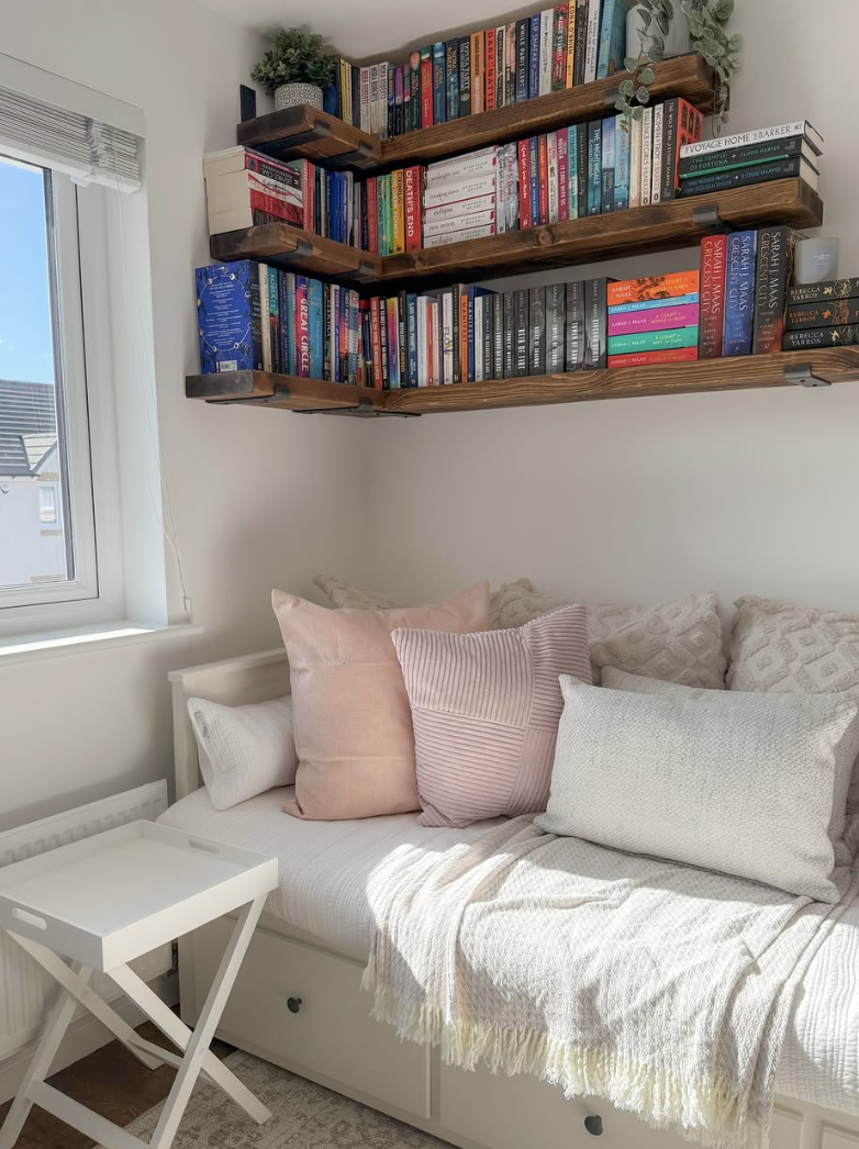 Bright reading nook with pink pillows, floating corner bookshelves, and white daybed