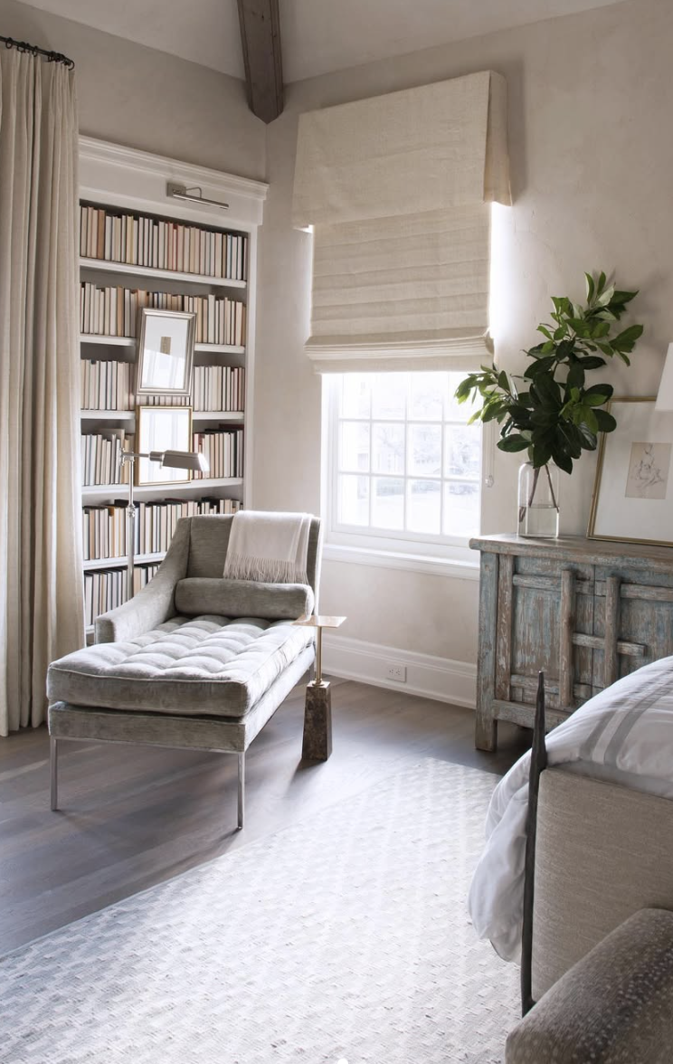 Minimalist reading nook with a velvet chaise lounge, neutral tones, wall-mounted bookshelves, and soft roman shades