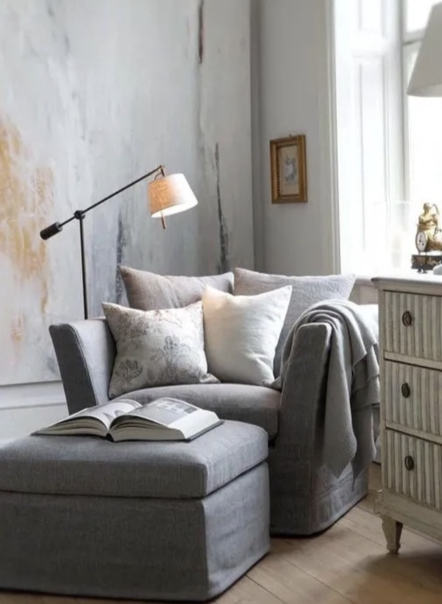 Grey armchair with ottoman and neutral pillows beside a large window with soft lighting and a floor lamp