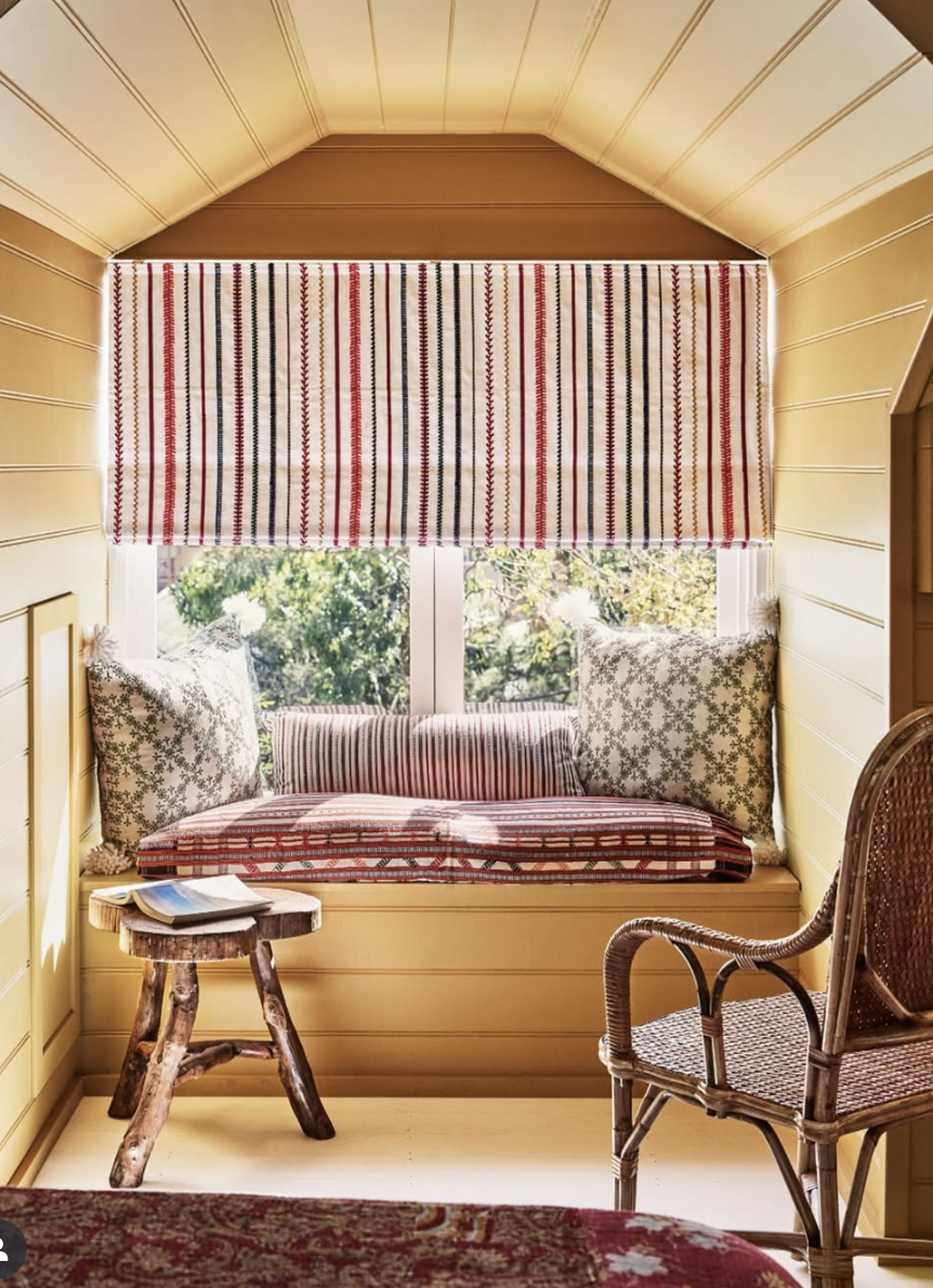 Colorful built-in window seat with red and white striped shades, patterned pillows, and rustic wood stool