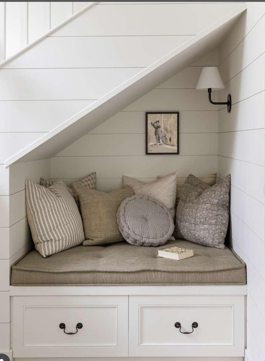 Neutral-toned reading nook built under a staircase with shiplap walls, plush cushions, and a wall sconce for lighting