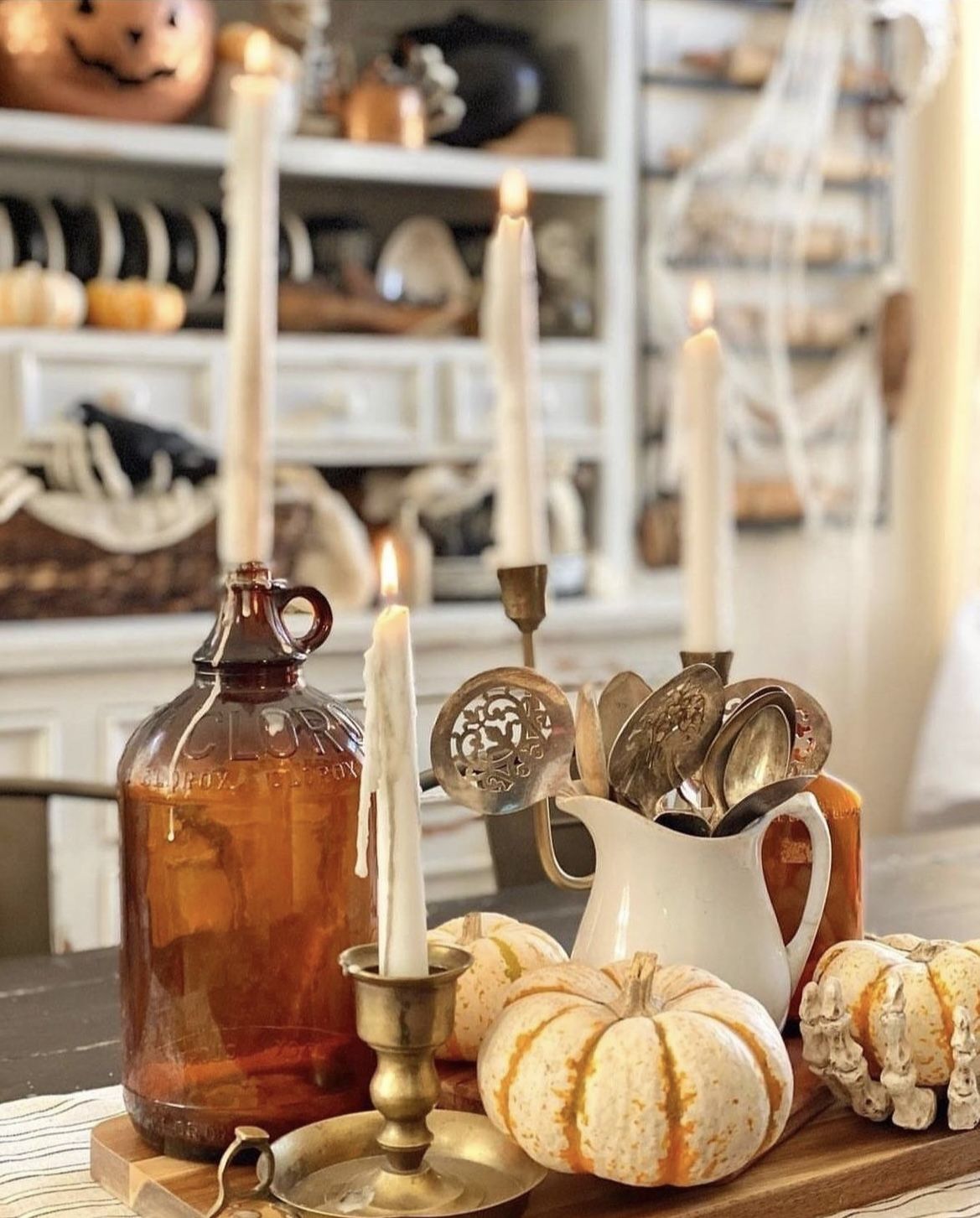 Rustic farmhouse fall centerpiece with mini pumpkins, taper candles, amber glass jug, brass candlesticks, and vintage silverware in a white pitcher.
