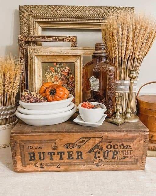 Rustic farmhouse fall vignette with a vintage butter crate, dried wheat, mini pumpkins, stacked plates, and antique frames.