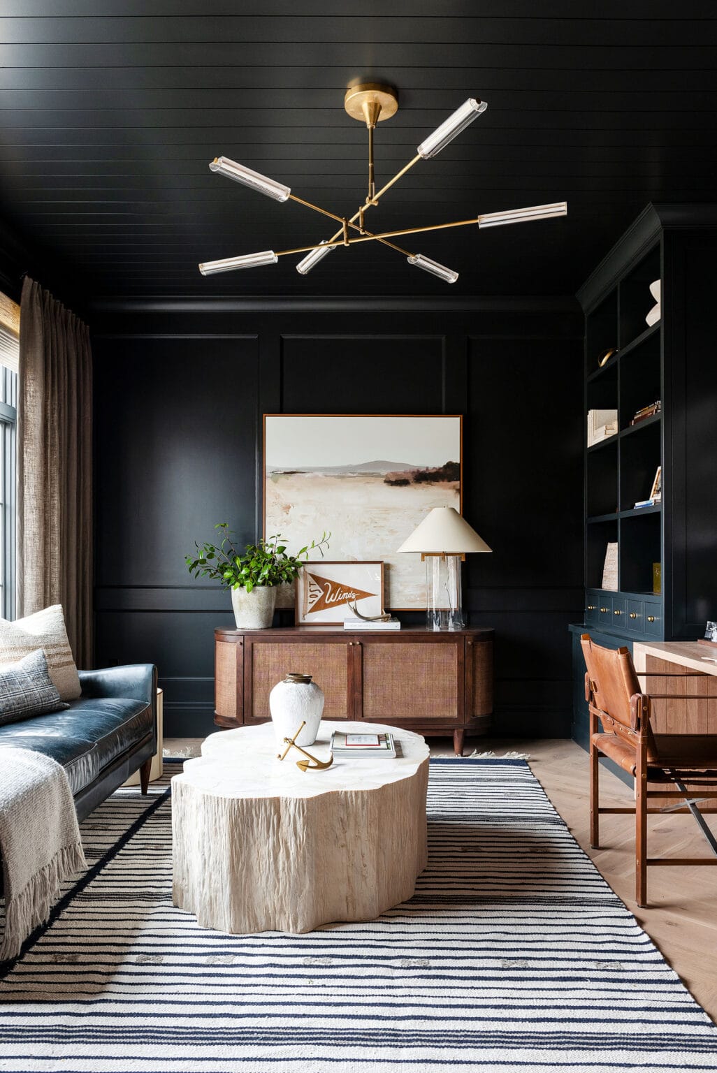 Studio McGee office featuring dark paneled walls, built-in shelving, a striped rug, and a brass modern chandelier.