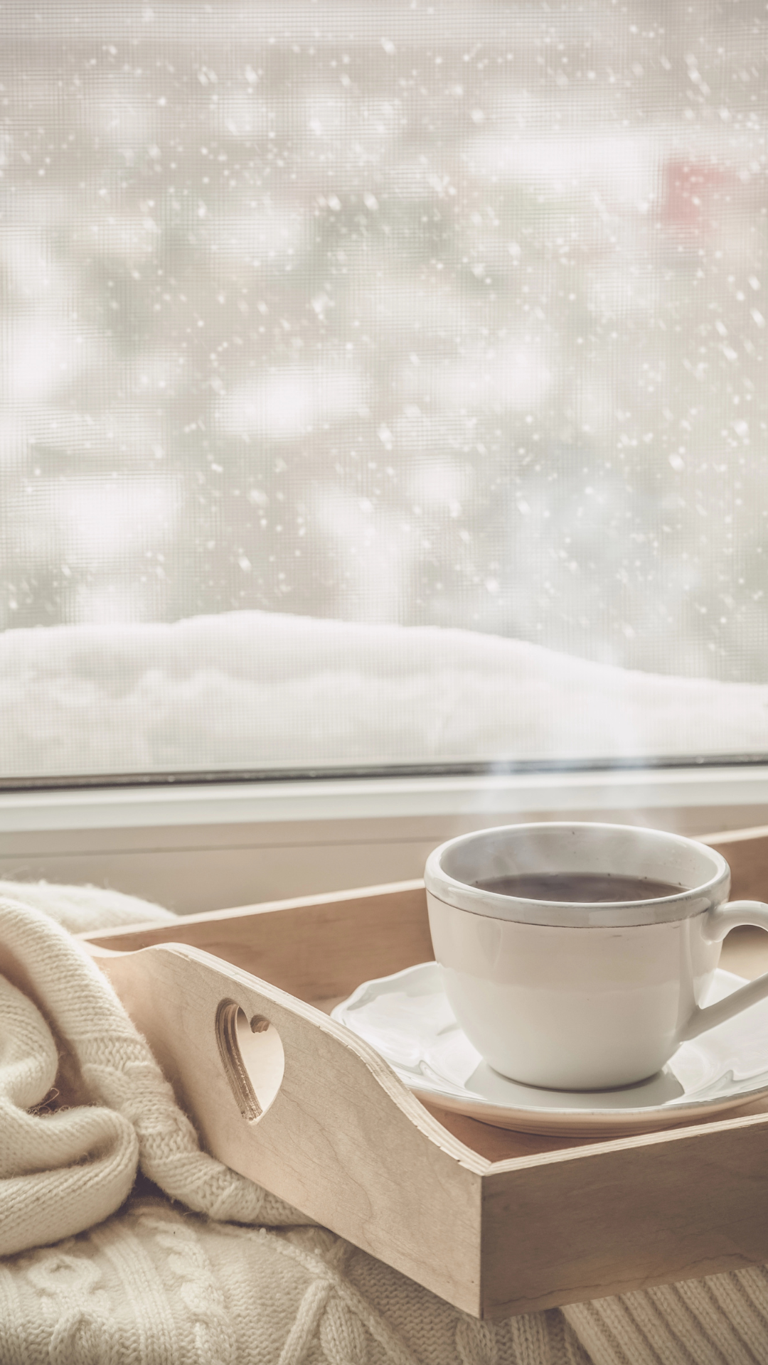 Cozy winter aesthetic wallpaper with steaming coffee on a wooden tray, knit blankets, and snow falling outside the window.