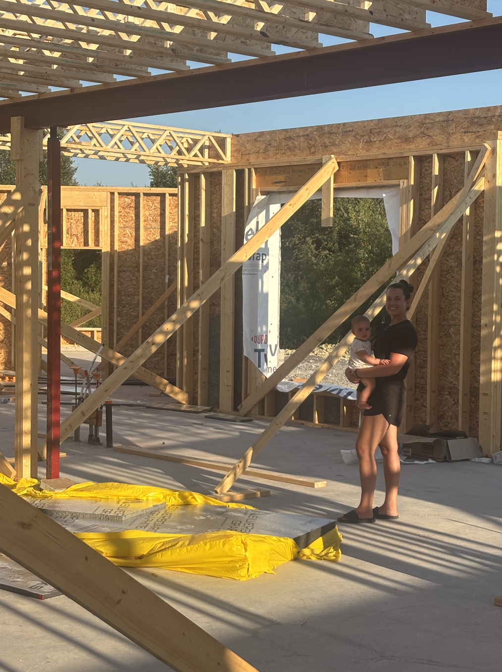 Mom holding baby inside a modern farmhouse during the framing stage, surrounded by exposed wooden beams and open wall structures.