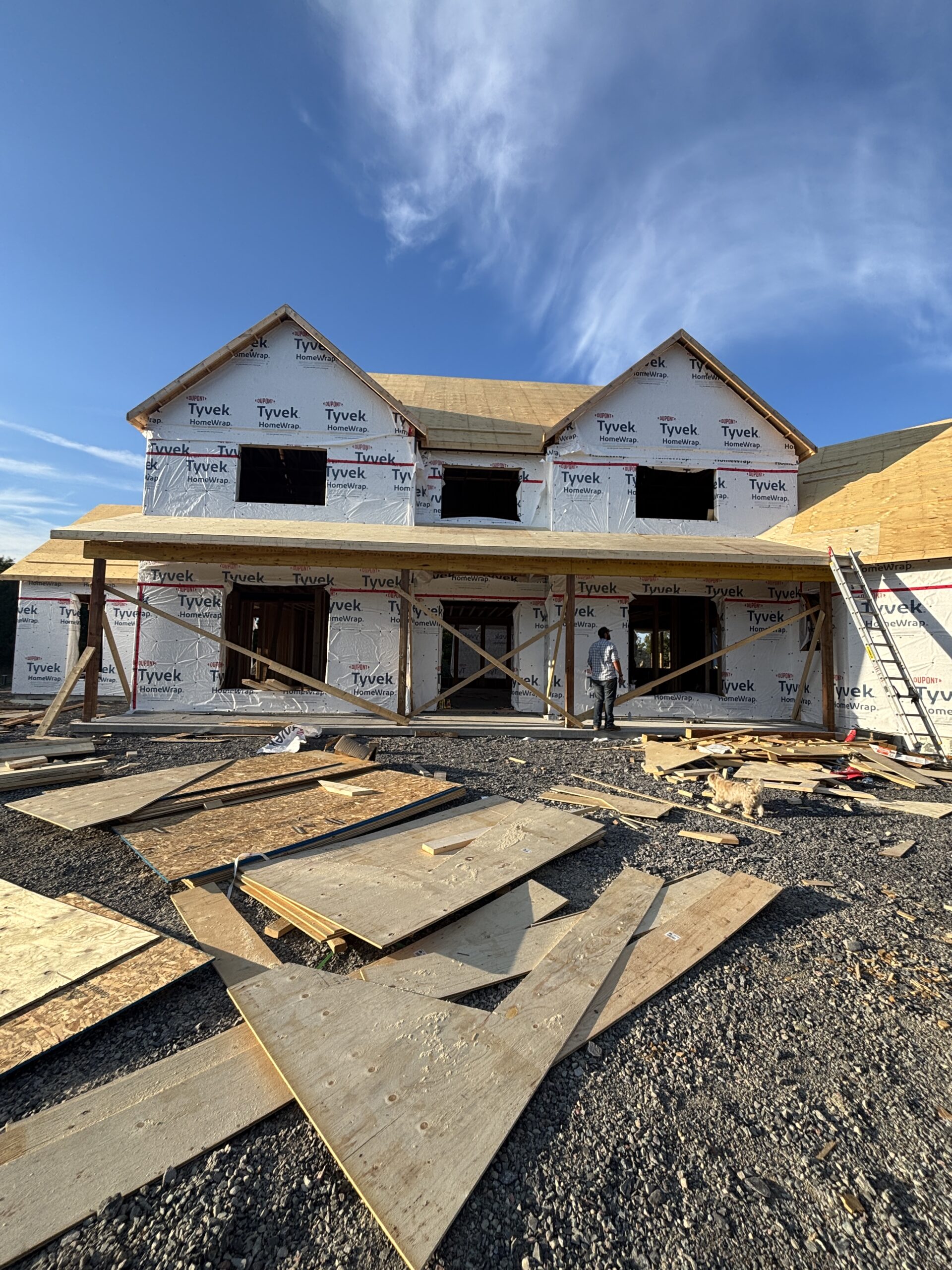 Front exterior of a modern farmhouse under construction with Tyvek wrap, showing framed walls, roof sheathing, and open window cutouts during the framing stage.