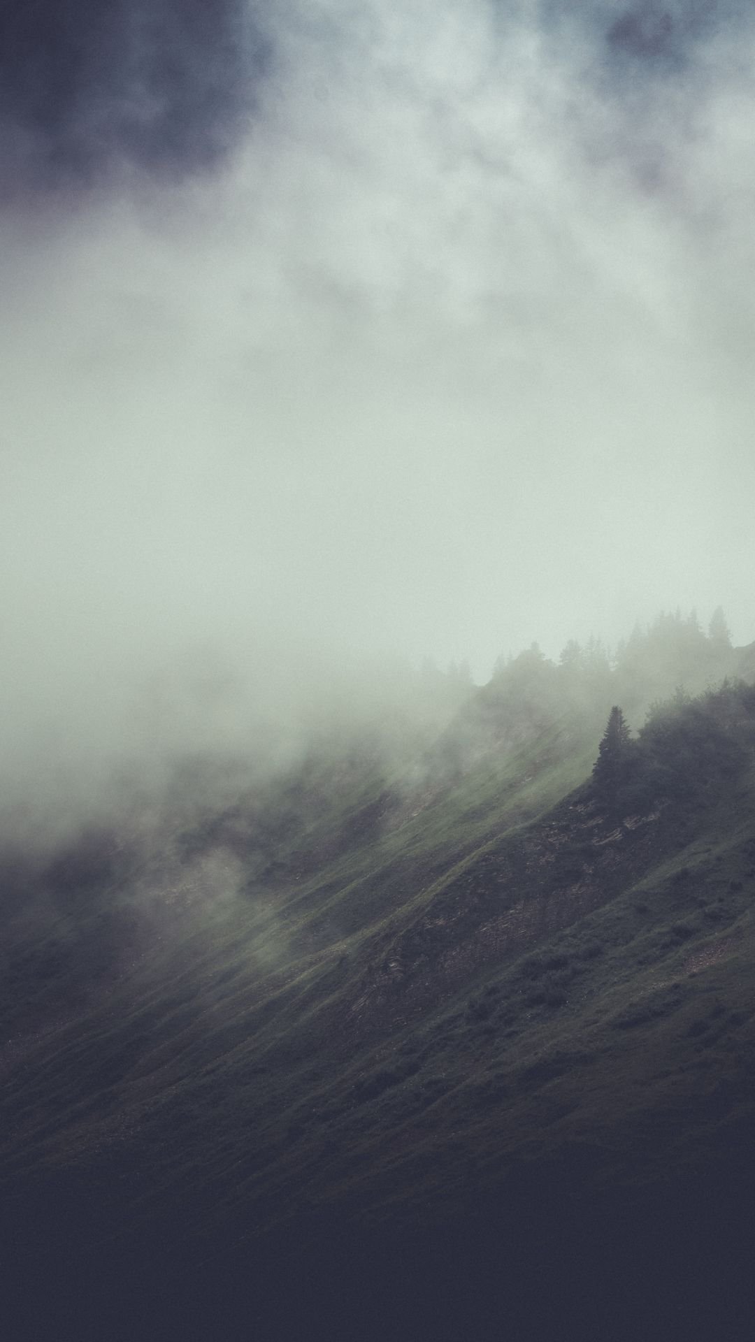 A minimalist moody winter wallpaper showing fog rolling over a green mountain with dark winter skies.