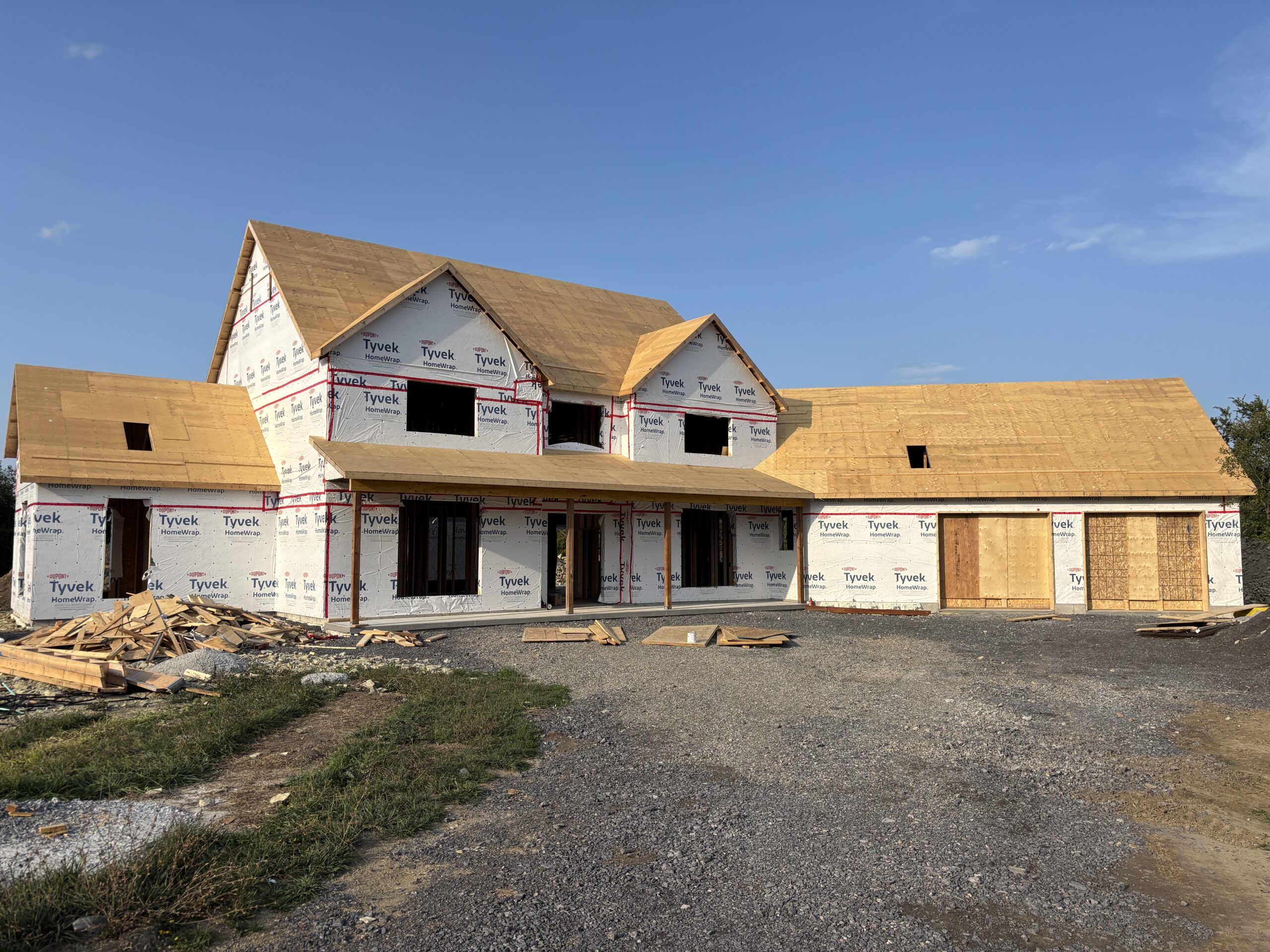 Front exterior of a modern farmhouse under construction, showing completed framing, Tyvek wrap, and roof sheathing during the house build process.