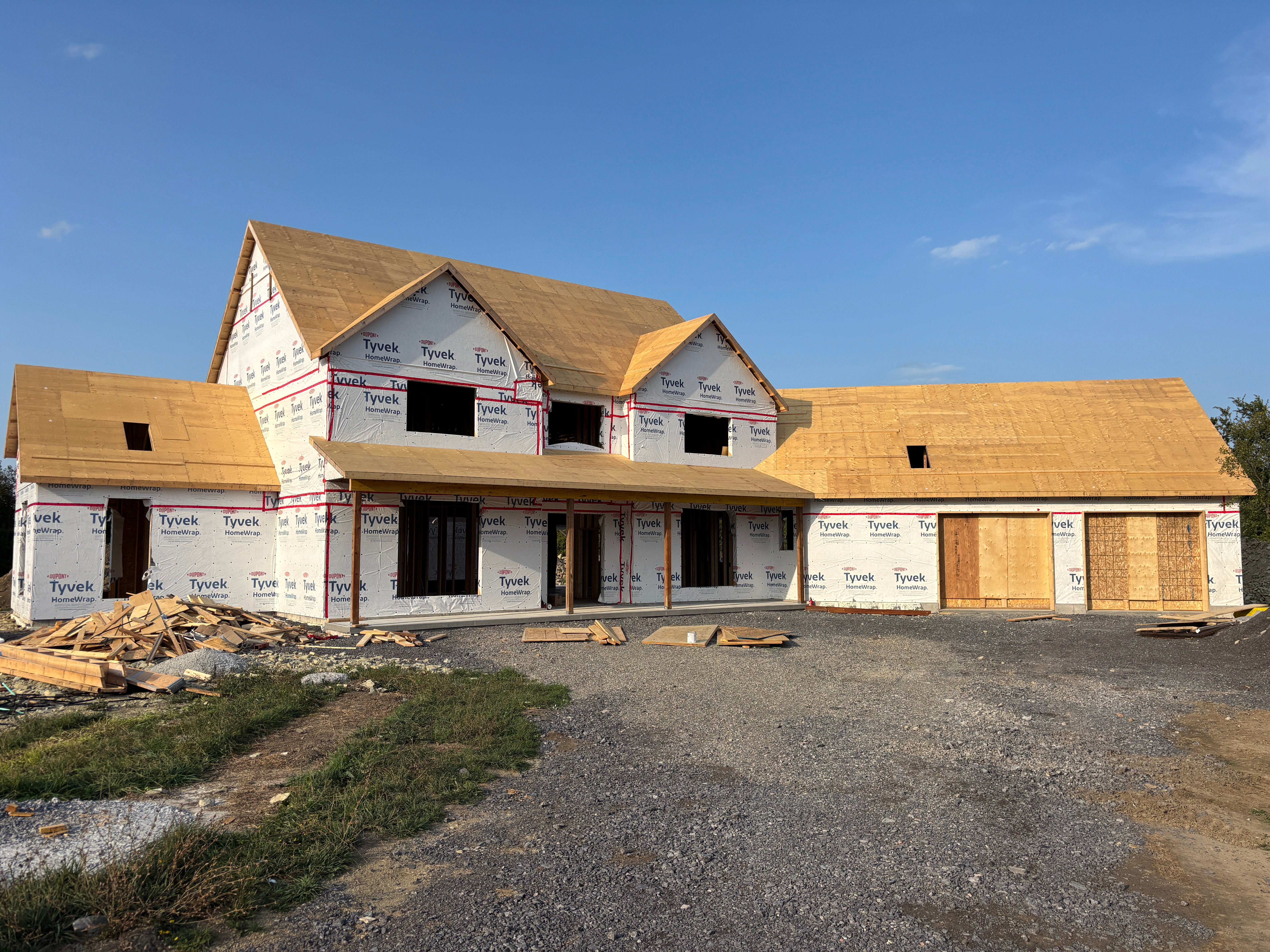Front exterior of a modern farmhouse under construction, showing completed framing, Tyvek wrap, and roof sheathing during the house build process.