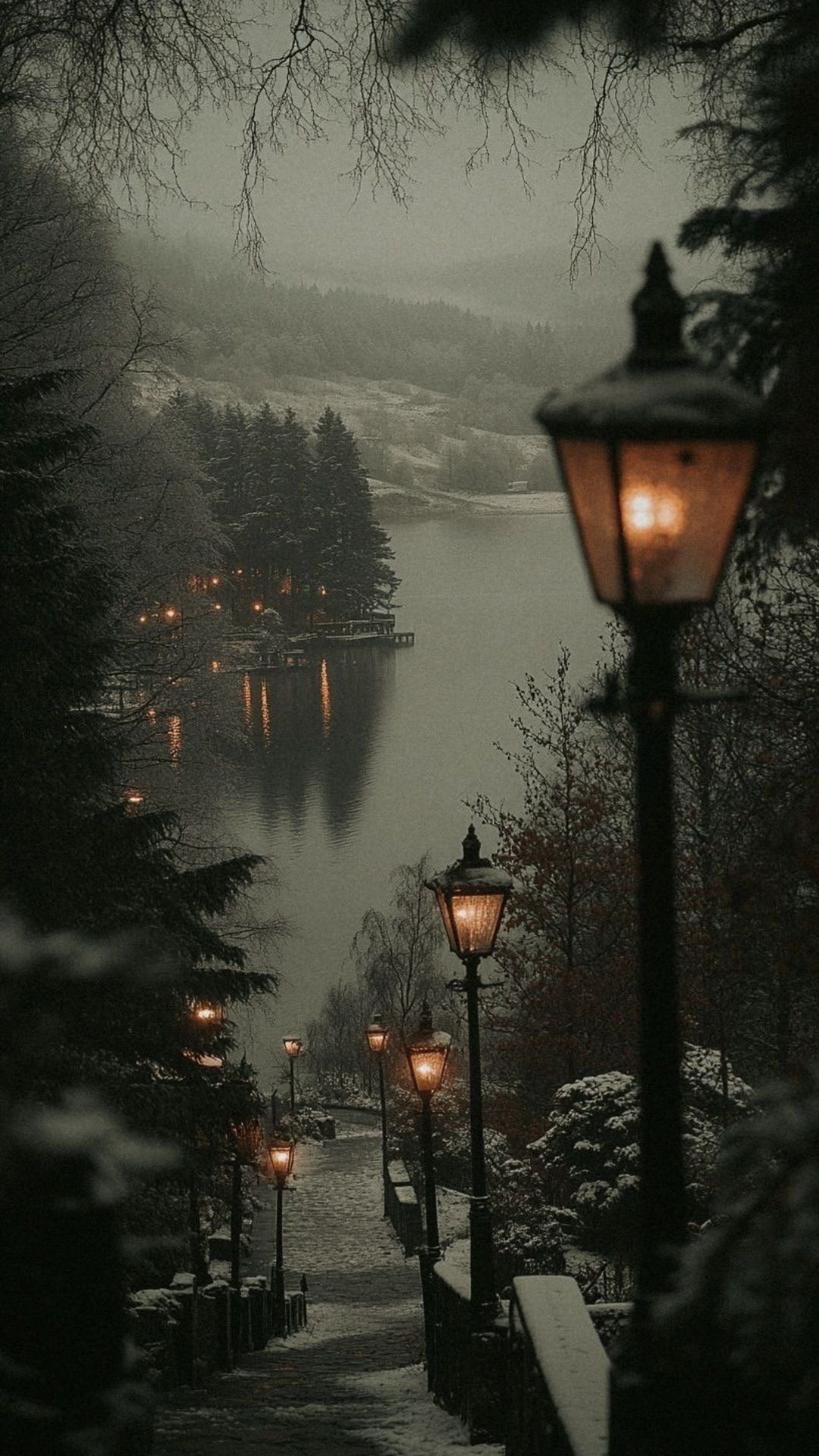 A moody winter iPhone wallpaper of a snow-covered path lined with lanterns leading to a foggy lake.